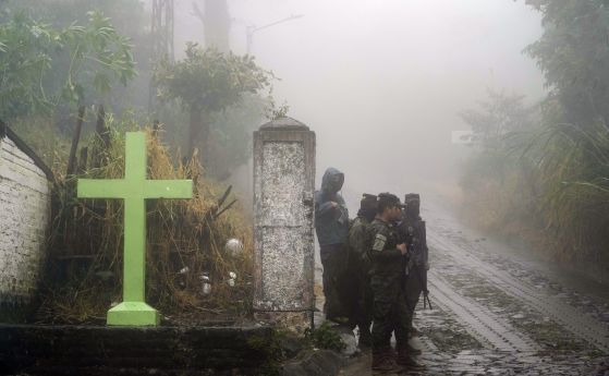 Войници стоят на стража под дъжда близо до кръст, който украсява пътя в Комасагуа, Салвадор, в понеделник, 10 октомври 2022 г. Петима войници загинаха в понеделник наблизо, когато стената на къща се срутила.