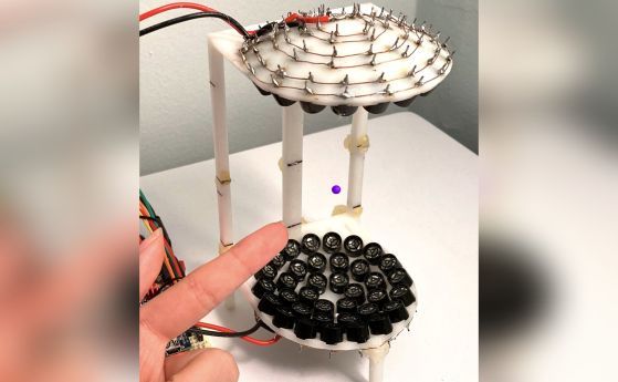 По-горе, мънисто (лилаво) е окачено във въздуха от звукови вълни, излъчвани от (черни) кръгли високоговорители, разположени в рамка с височина шест инча, отпечатана на 3D принтер.