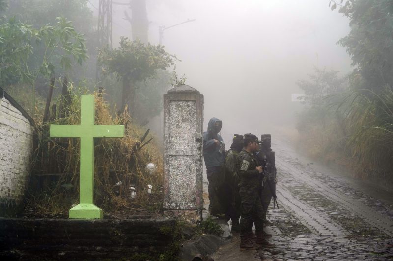 Войници стоят на стража под дъжда близо до кръст, който украсява пътя в Комасагуа, Салвадор, в понеделник, 10 октомври 2022 г. Петима войници загинаха в понеделник наблизо, когато стената на къща се срутила.