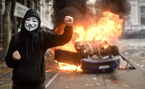 Протестиращ с маска Anonymous пред запалена кола